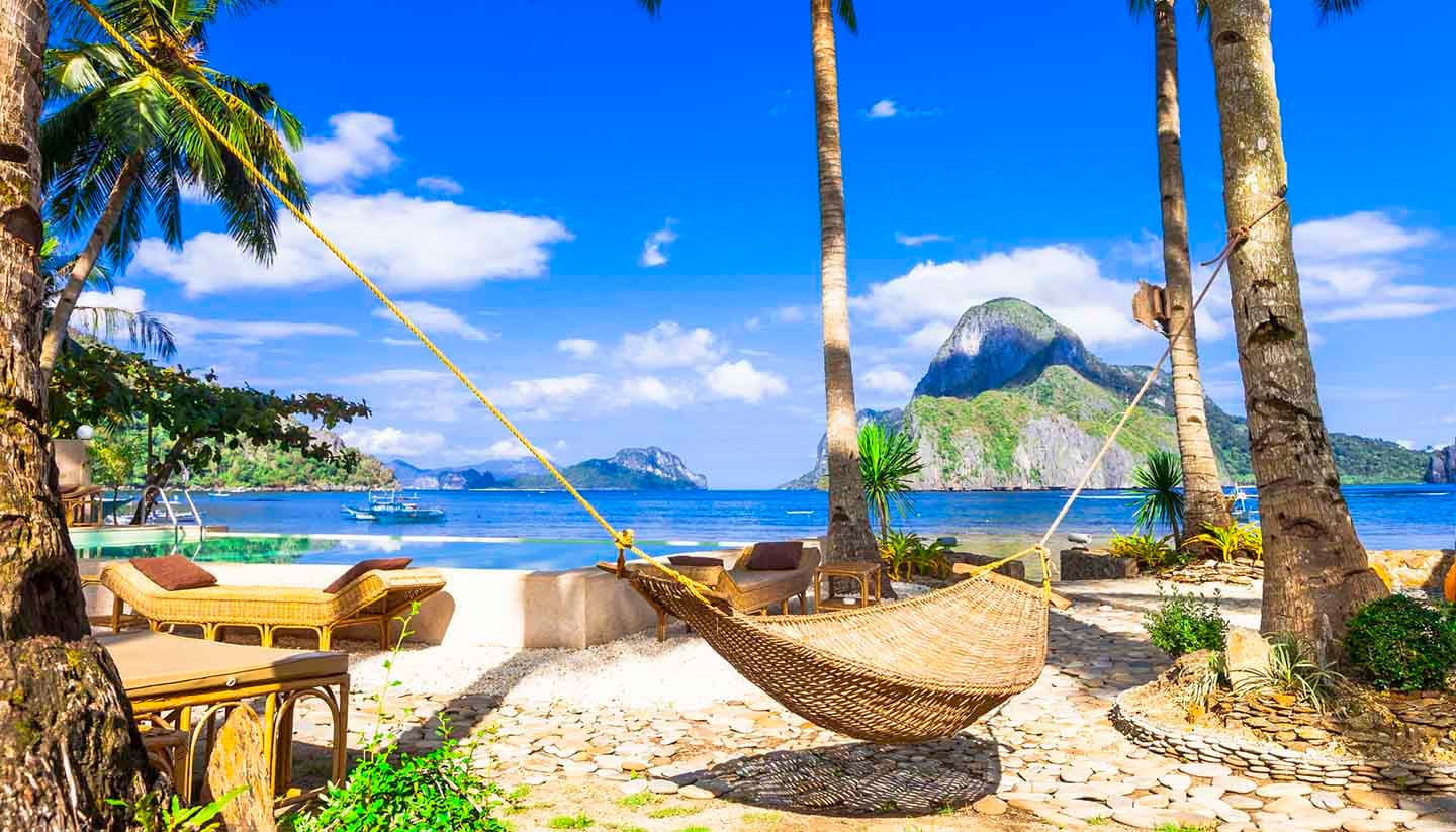 A woven hammock strung between palm trees on a beachfront terrace, looking out over a calm bay to limestone islands.